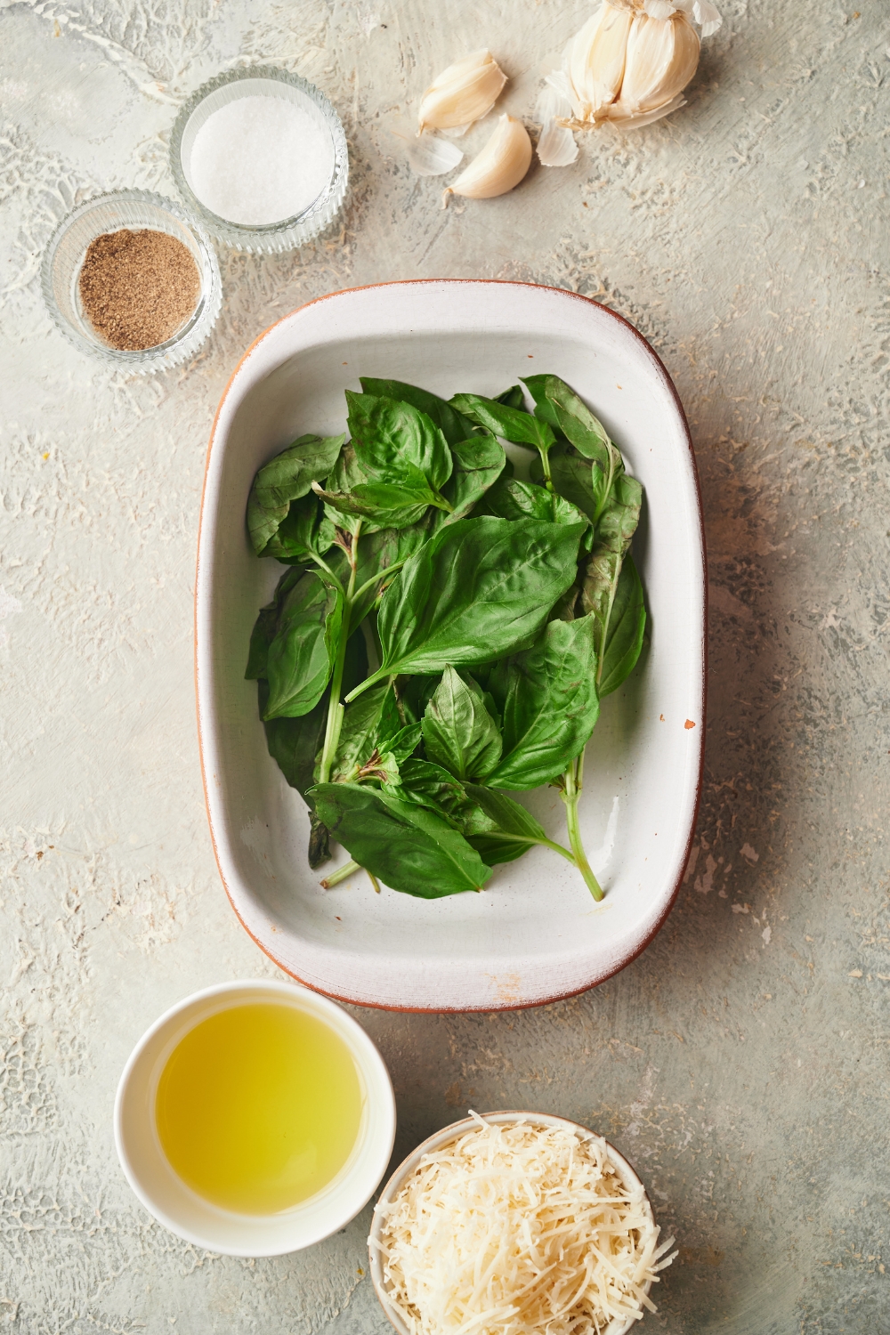 An assortment of ingredients including a baking dish filled with fresh basil, garlic cloves, and bowls of oil, parmesan cheese, salt, and pepper.