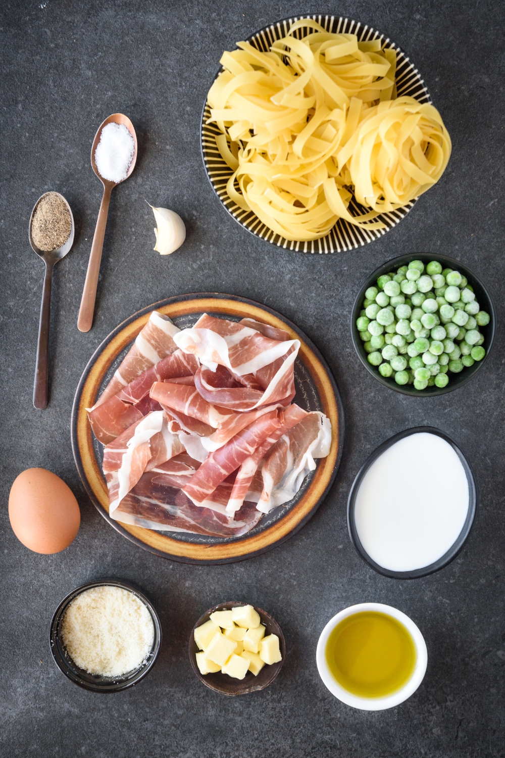 An assortment of ingredients including a plate of prosciutto and bowls of dried pasta, frozen peas, parmesan cheese, butter cubes, oil, cream, an egg, and spoonfuls of salt and pepper.