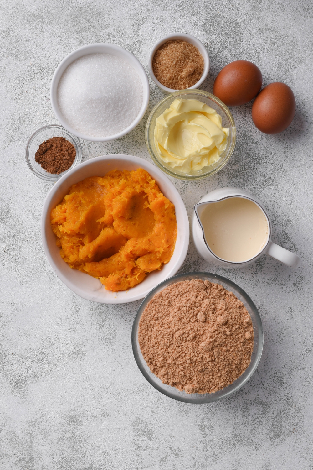 An assortment of ingredients including bowls of graham cracker crumbs, milk, pumpkin puree, butter, sugar, brown sugar, spices, and two eggs, all on a grey counter.