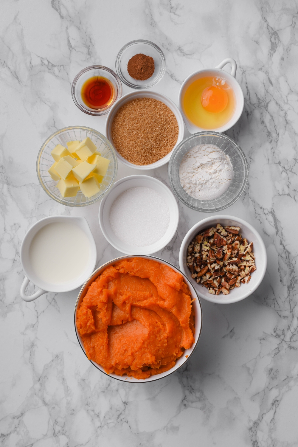 An assortment of ingredients including bowls of sweet potato puree, chopped pecans, brown sugar, vanilla extract, milk, sugar, butter cubes, an egg, and spices.