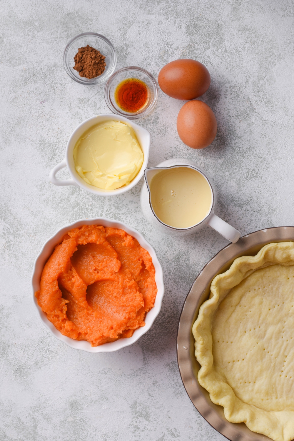 An assortment of ingredients including a pie pan with a pie crust in it and bowls of sweet potato puree, butter, vanilla extract, spices, condensed milk, and two eggs, all on a grey counter.