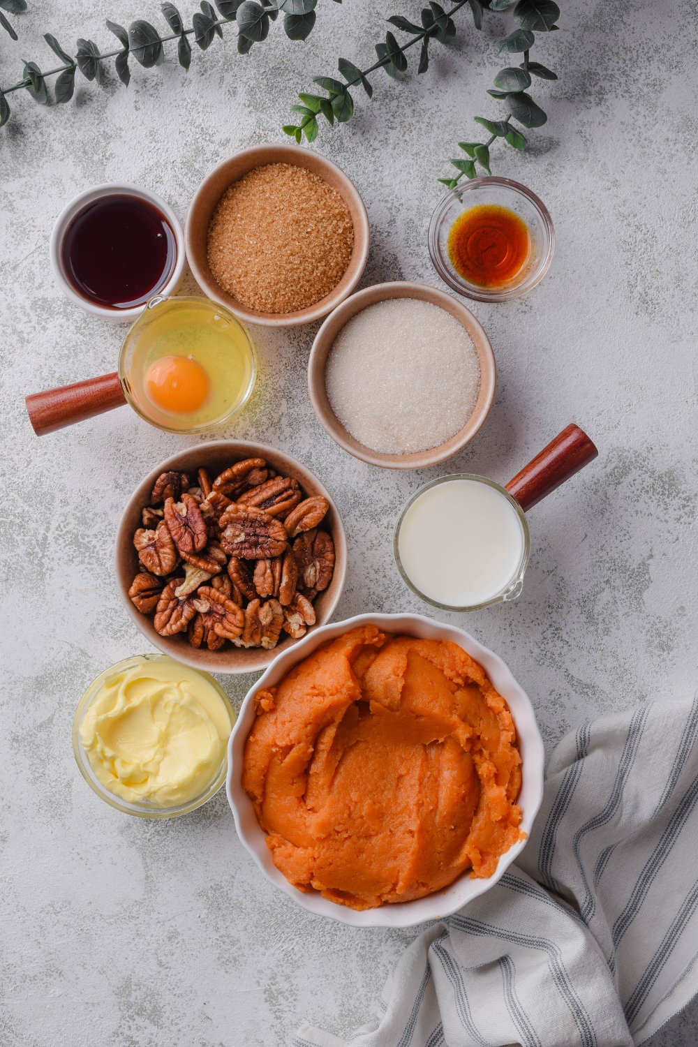 An assortment of ingredients including bowls of pecans, sweet potato puree, brown sugar, white sugar, vanilla extract, an egg, butter, and milk, all on a grey counter.