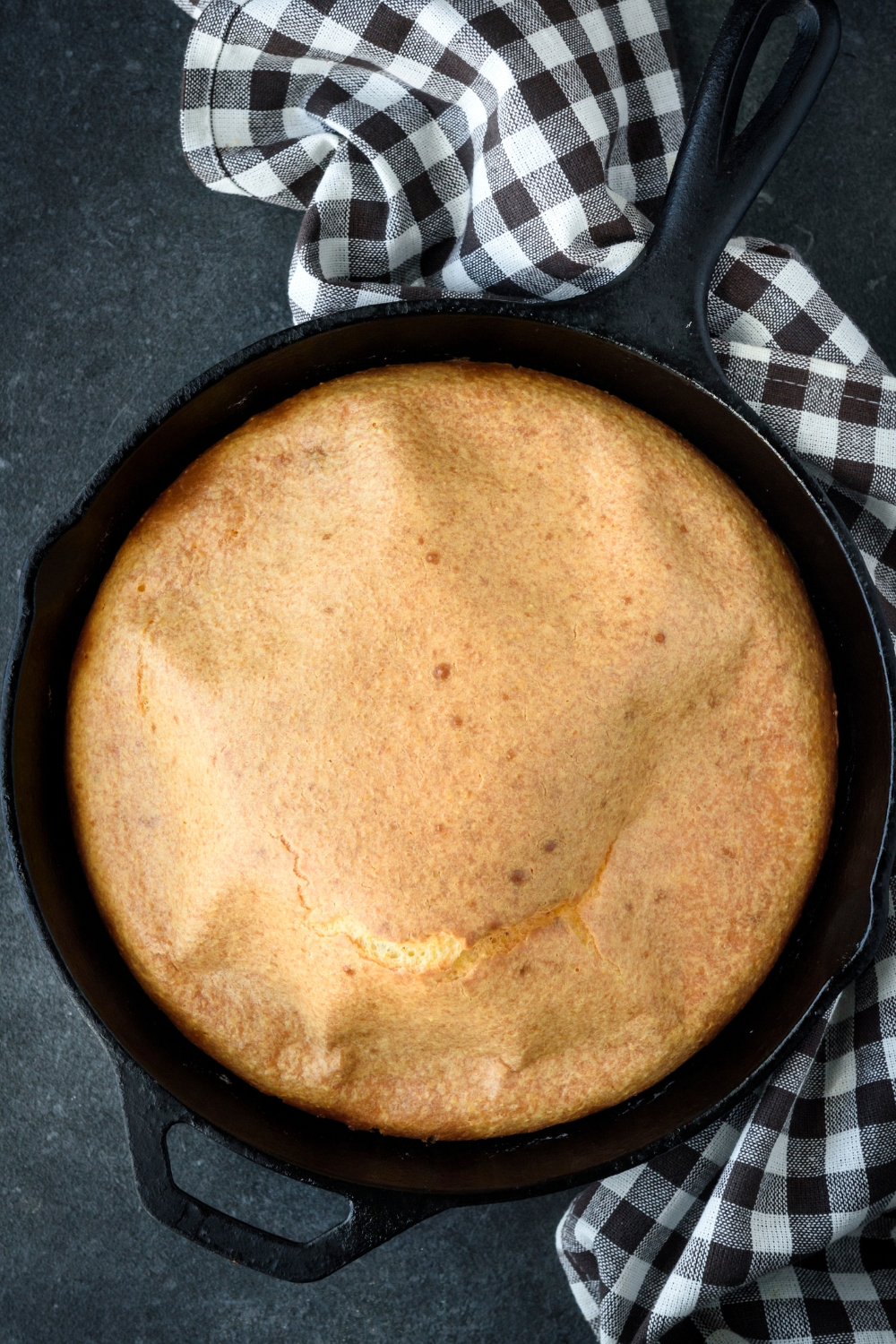 A cast iron skillet full of golden brown cornbread on a black counter. A black and white dish towel is under the skillet.