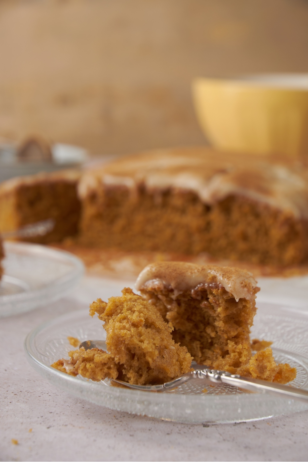 A piece of pumpkin cake on a fork on a clear glass plate. The rest of the pumpkin cake is in the background.