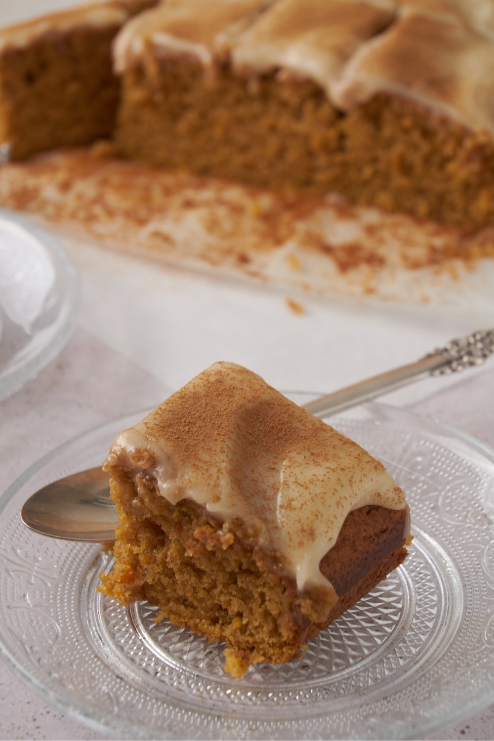 A piece of pumpkin cake on a glass plate with a silver spoon behind it. The rest of the pumpkin cake is in the background.