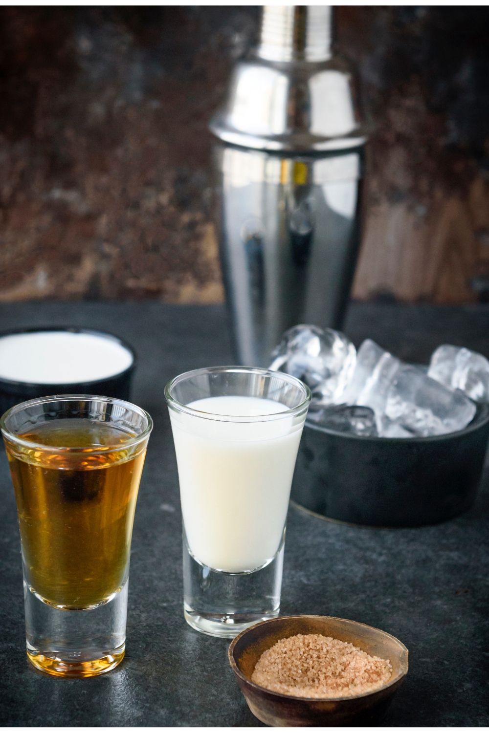 A metal shaker, ice, a shot glass full of Rumchata, a shot glass of fireball, and a small bowl of cinnamon and sugar mixture sit on a black countertop.