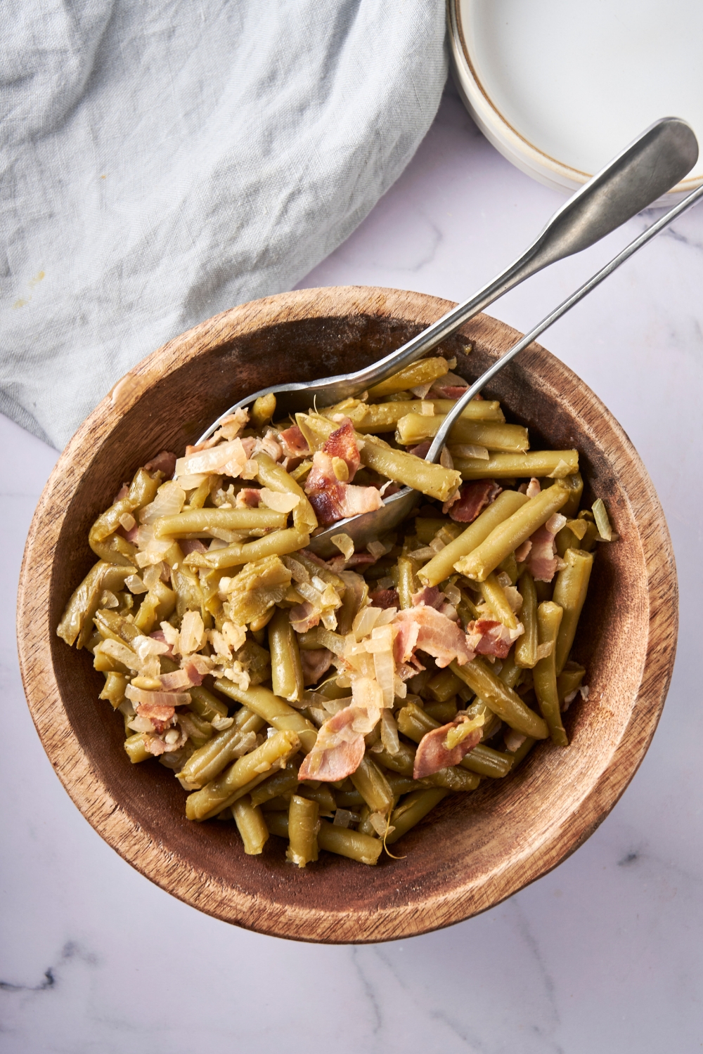 A large wooden bowl full of green beans and bacon on a white counter. Serving fork and spoon are resting on the bowl.
