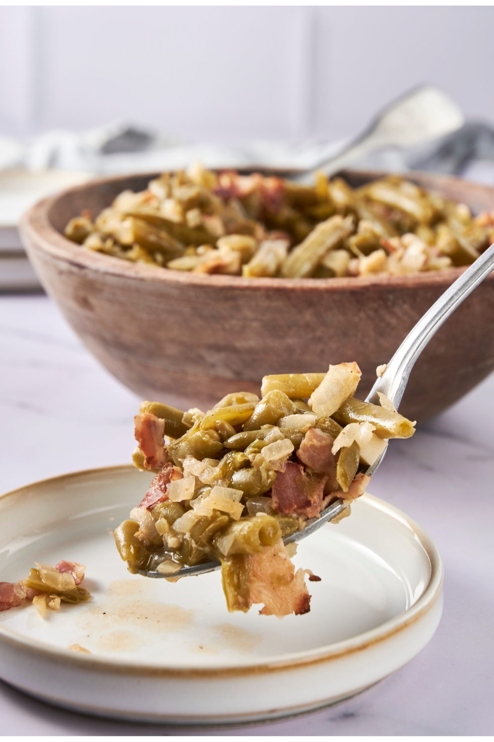 A serving spoon scoops green beans onto a white plate. A large wooden bowl of beans is in the background.