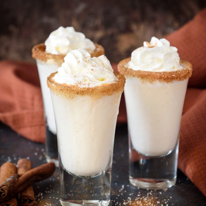 Three cinnamon toast crunch shots rimmed with cinnamon and sugar and topped with whipped cream on a black counter top.