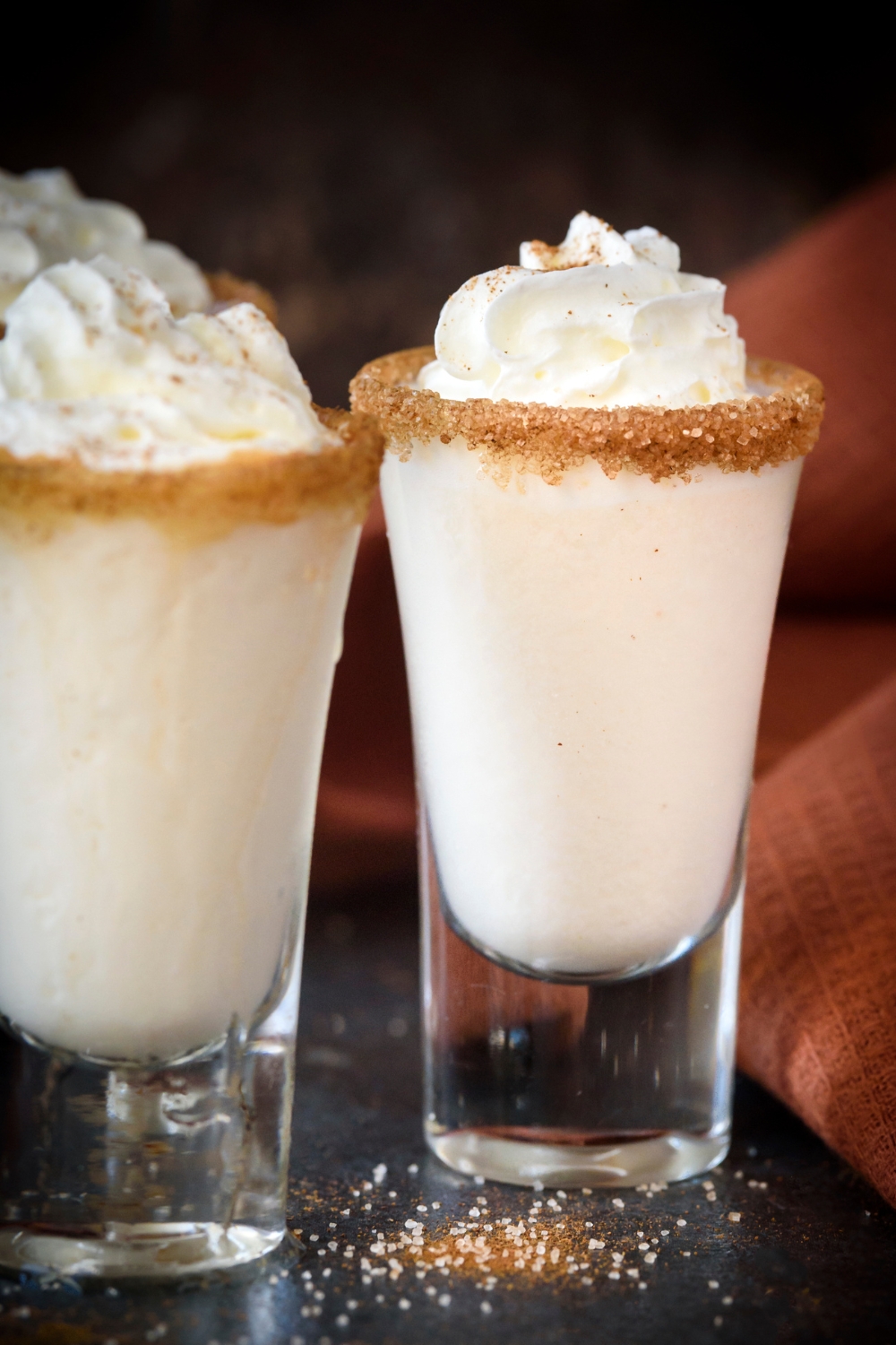 Three cinnamon toast crunch shots rimmed with cinnamon and sugar topped with whipped cream on a black counter top.