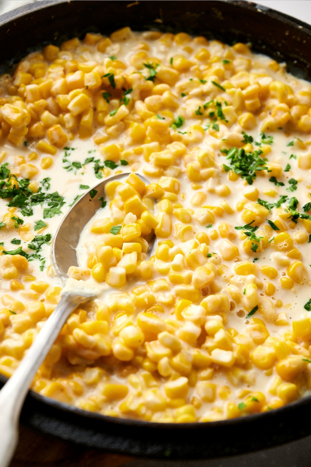 Creamed corn in a black pot with a silver spoon. The corn is garnished with fresh herbs.