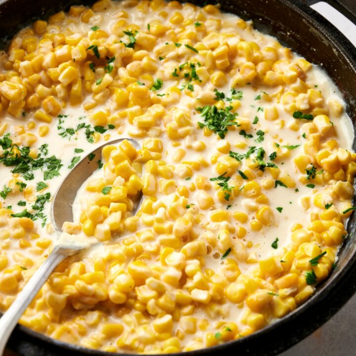 Creamed corn in a black cast iron pot garnished with fresh herbs. A spoon is in the pot.