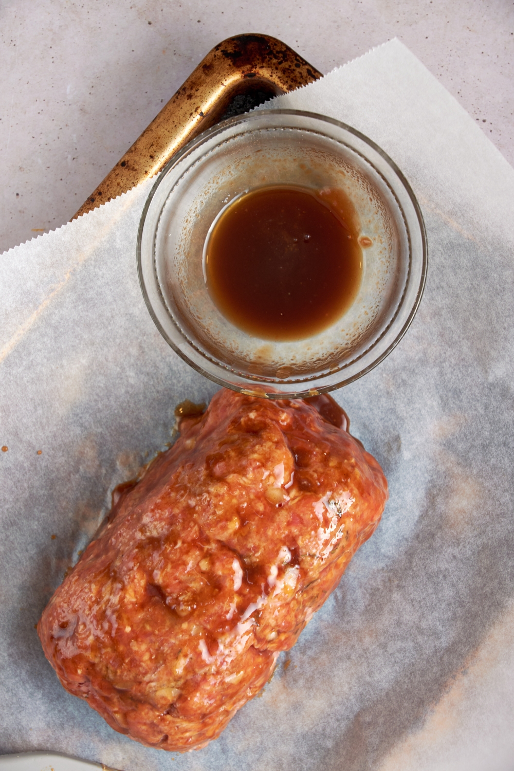 An uncooked meatloaf with barbecue sauce spread over it. A glass bowl of barbecue sauce is next to it.