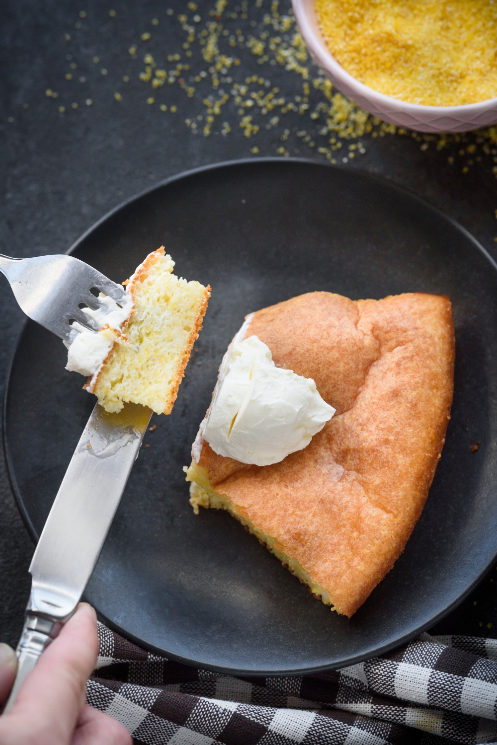 Someone cuts a bite of cornbread out of a slice on a black plate. The cornbread is topped with cream cheese.