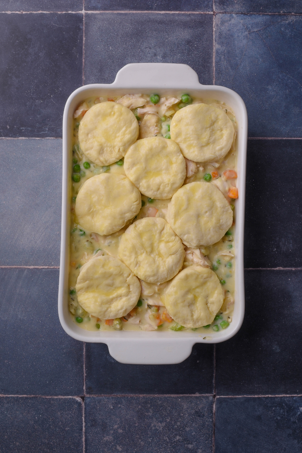 A white casserole dish of chicken pot pie filling and eight uncooked round biscuits on top.