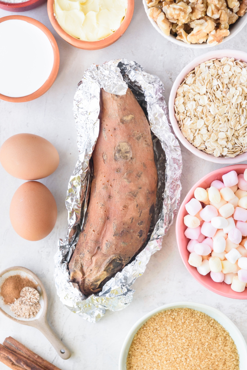 One large sweet potato partially wrapped in foil is on a white counter. Surrounding the potato are bowls of walnuts, oats, marshmallows, brown sugar, flour, cream, and two eggs.