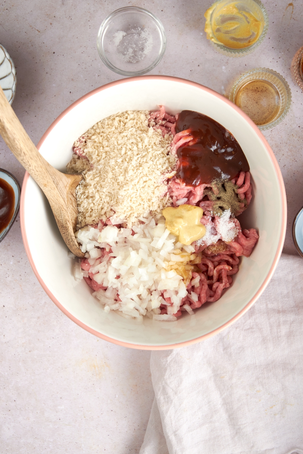 Ground beef, bread crumbs, mustard, chopped onion, and seasonings in a white mixing bowl with a wooden spoon. Empty ingredient bowls are nearby.