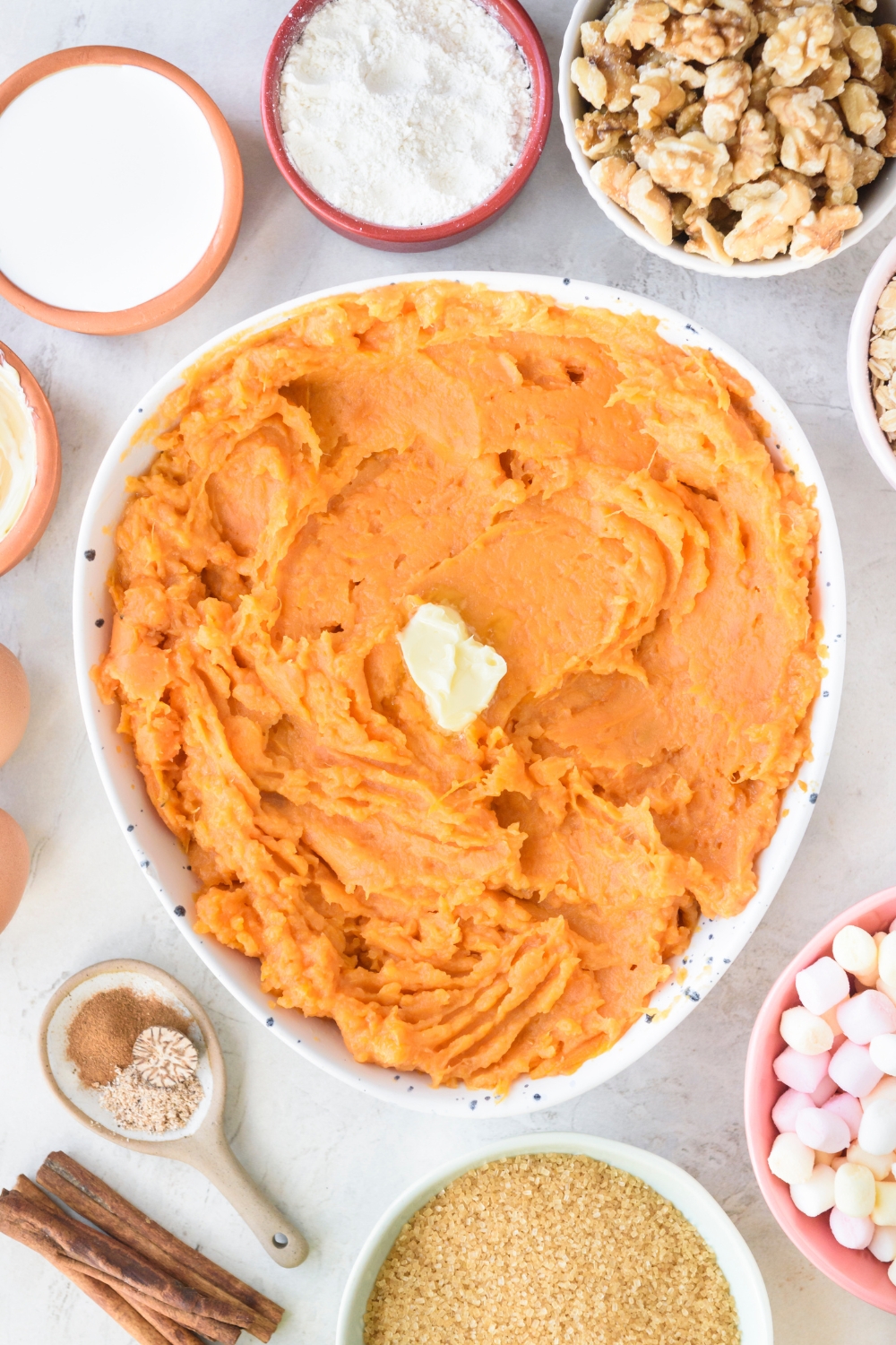 A large white bowl full of mashed sweet potatoes with a pat of butter in the middle. Bowls full of walnuts, marshmallows, brown sugar, spices, flour, and cream surround the large bowl