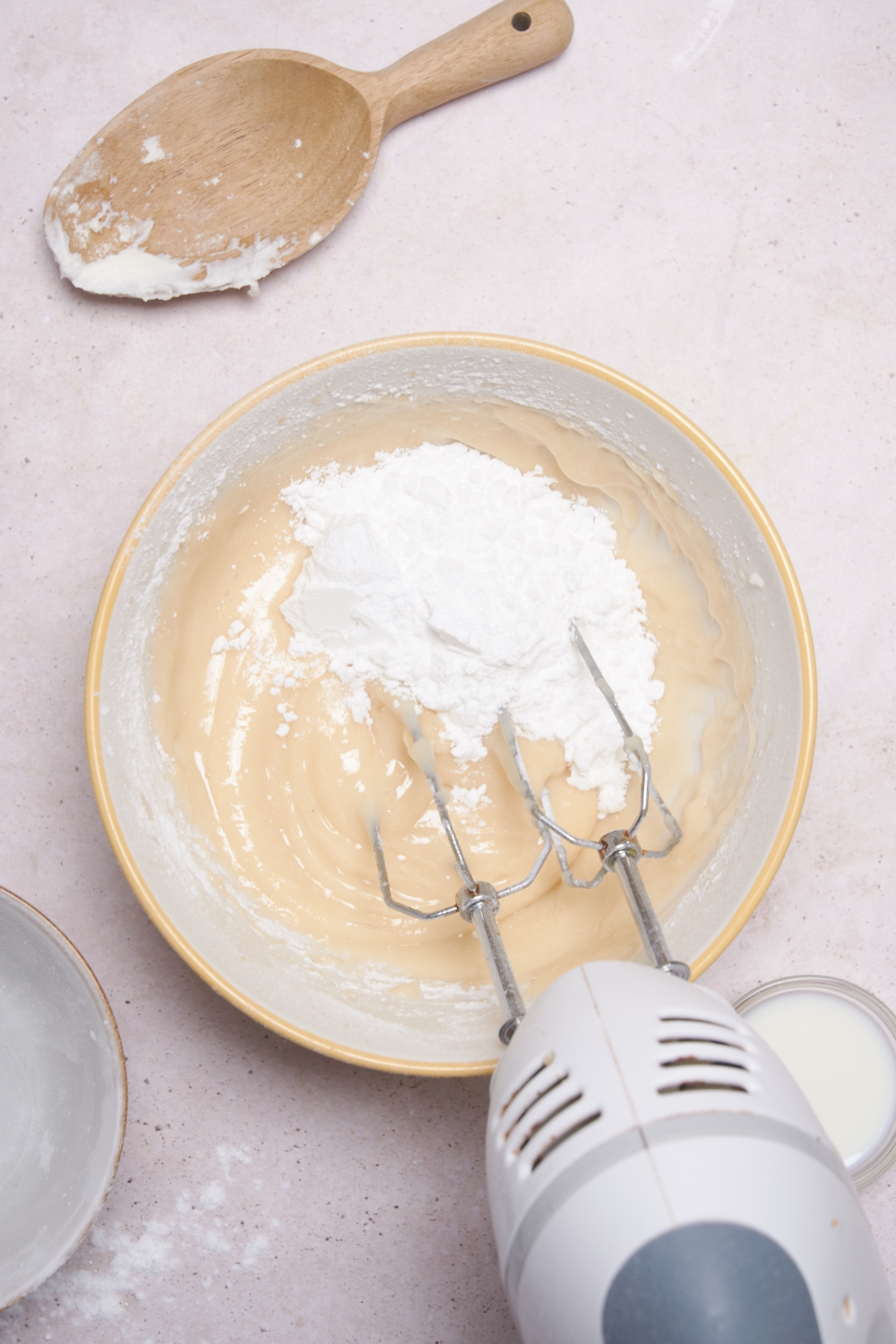 An electric mixer mixing the cream cheese frosting in a white mixing bowl. A wooden scoop with flour on it is nearby.