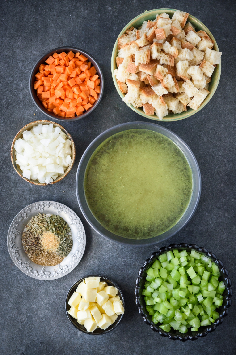 An assortment of ingredients including bowls of bread cubes, butter cubes, diced celery, diced carrots, diced onion, broth, and spices, all on a black counter.