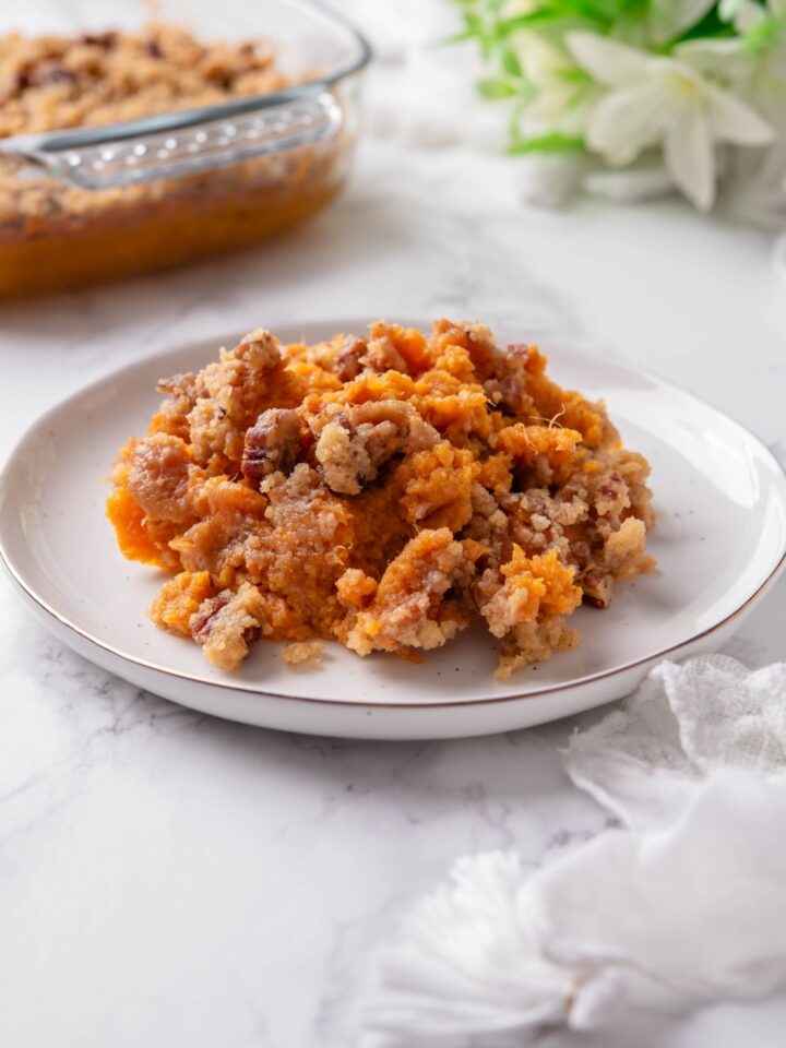 A plate filled with sweet potato casserole mixed with pecans and brown sugar. The rest of the casserole is in the background.