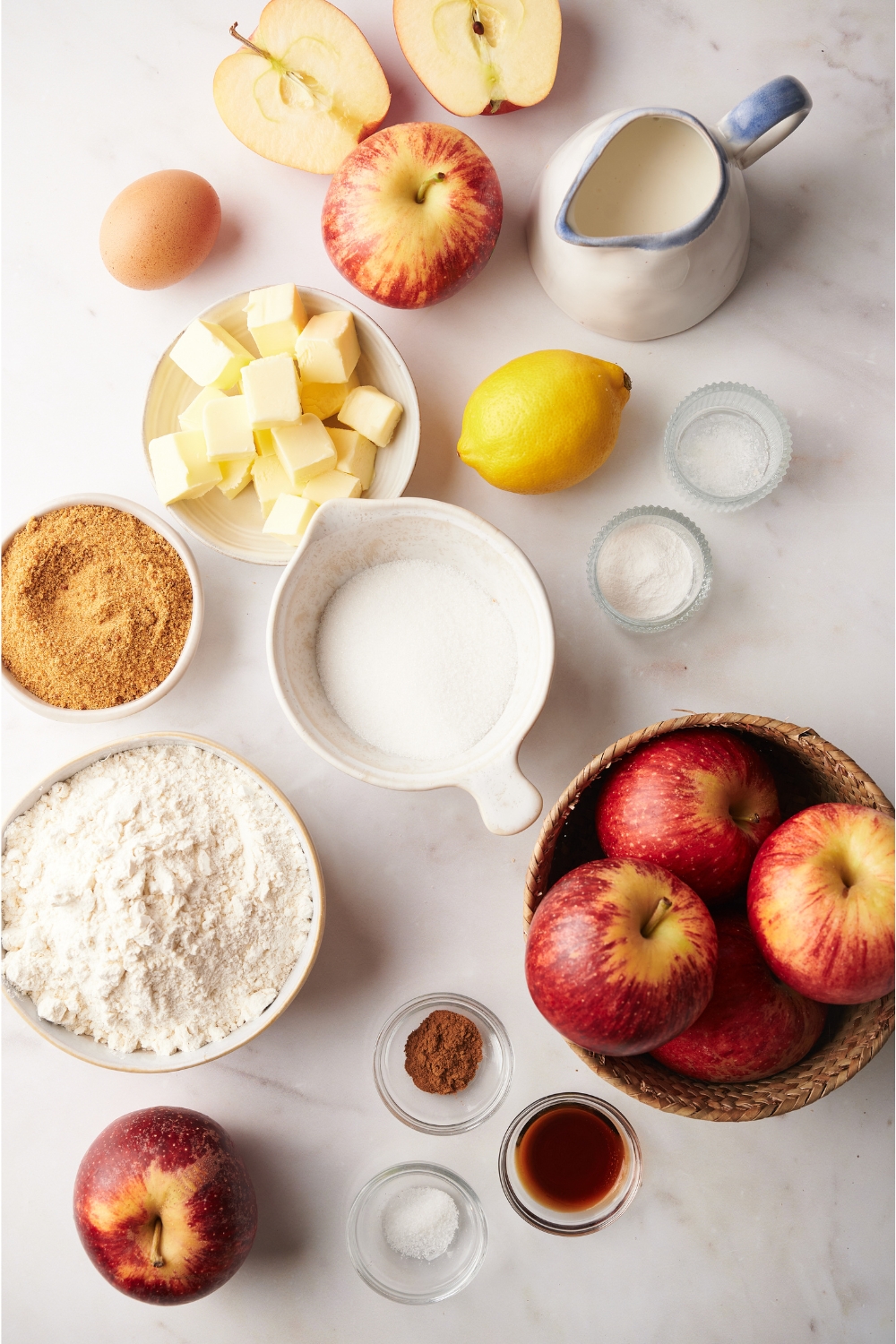 A countertop with apples, lemon, butter, an egg, brown sugar, white sugar, milk, and seasonings in various bowls.