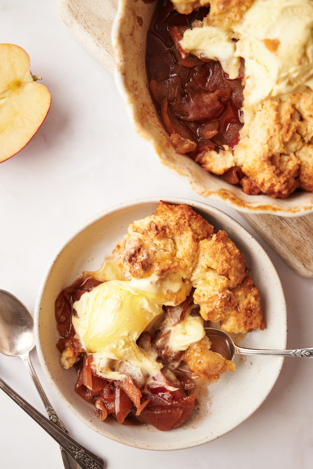 A plate with apple cobbler and a scoop of vanilla ice cream. The remaining cobbler is in a pie dish behind the plate on a wooden serving board.