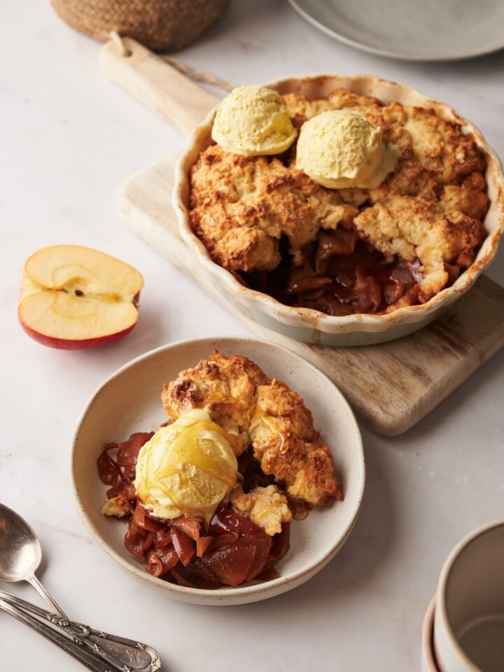 A plate with apple cobbler and a scoop of vanilla ice cream. The remaining cobbler is in a pie dish behind the plate on a wooden serving board.
