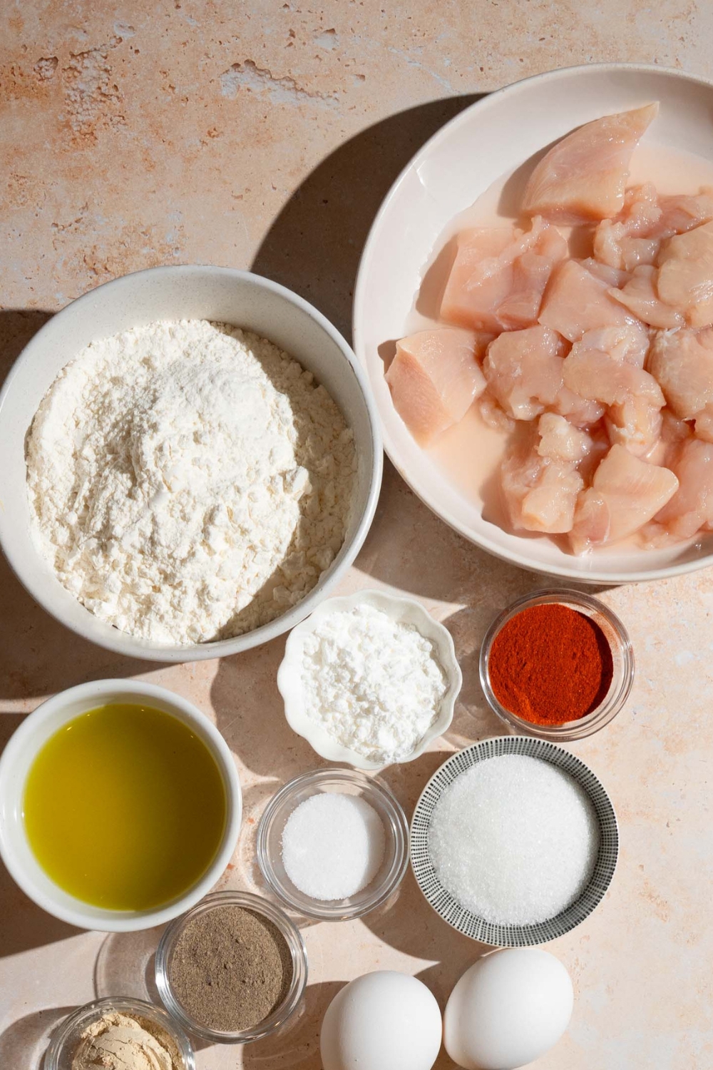 A bowl of chicken nuggets, a bowl of flour, a bowl of cornstarch, a bowl of paprika, a bowl of sugar, and a bowl of pickle juice all on a white counter.