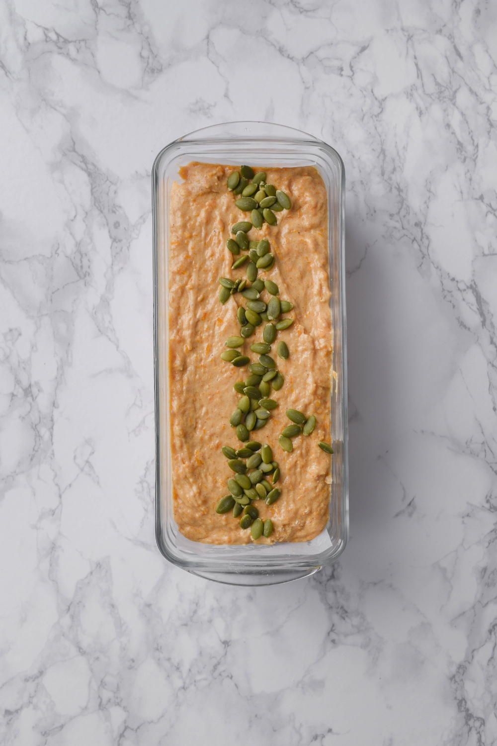 A loaf pan with pumpkin loaf batter topped with pumpkin seeds.