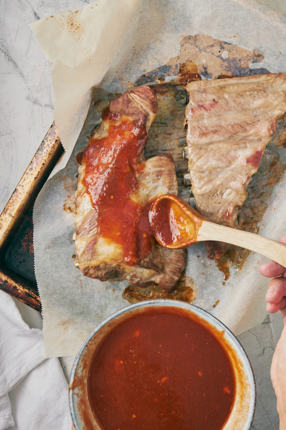 A hand pouring bbq sauce on top of riblets in a baking sheet lined with parchment paper.