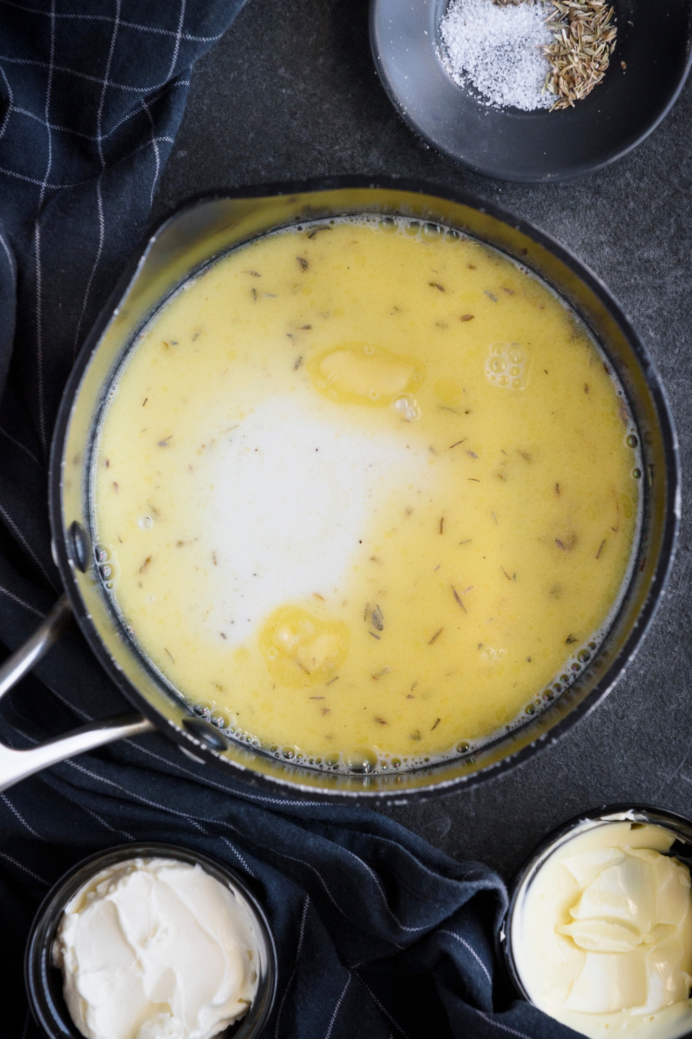 Milk, butter, and seasonings are in a silver sauce pan on a black counter.