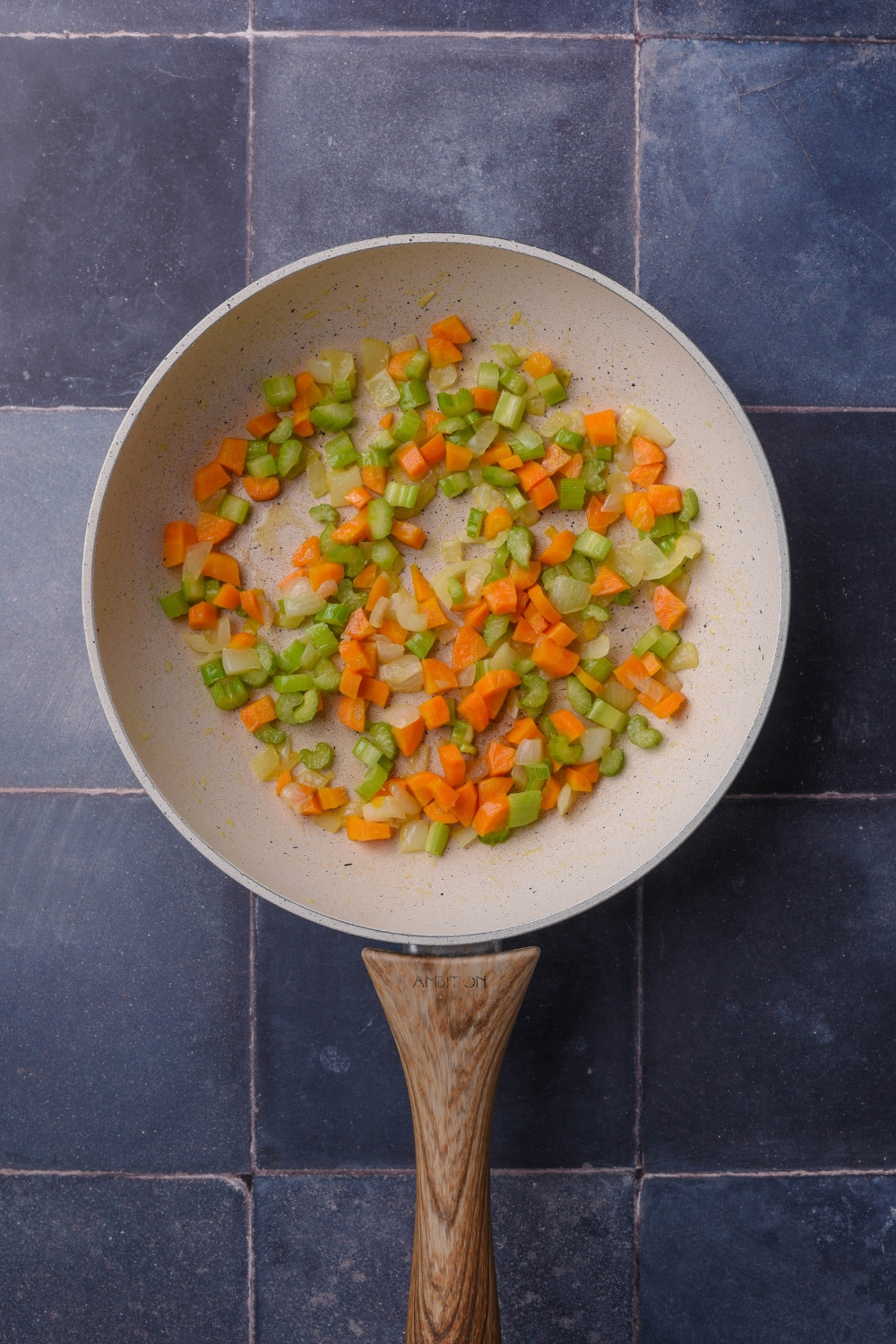 chopped carrots, celery, and onion sautéing in a white pan with a wooden handle.