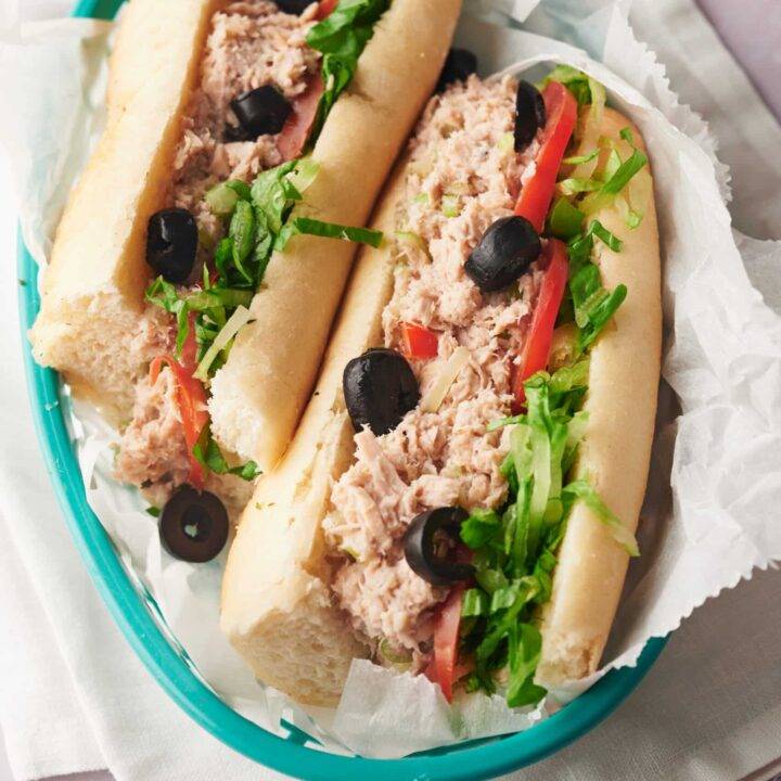Two halves of a tuna salad sandwich topped with olives in a blue basket lined with paper.