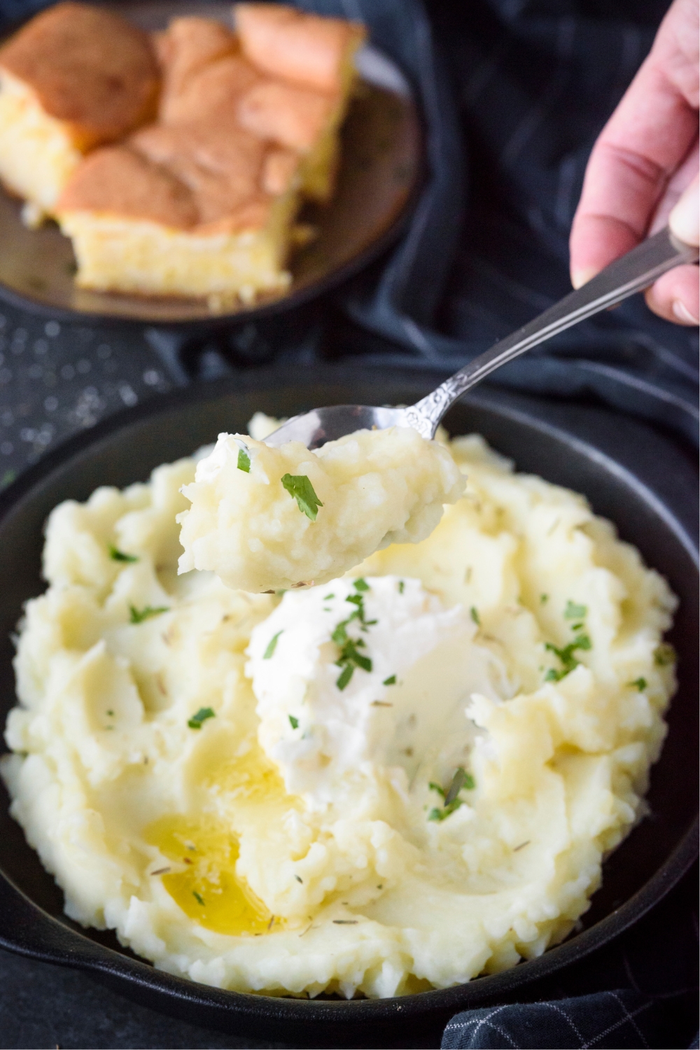 A hand holding a spoon scooping mashed potatoes out of a black bowl. The potatoes are topped with sour cream and butter.