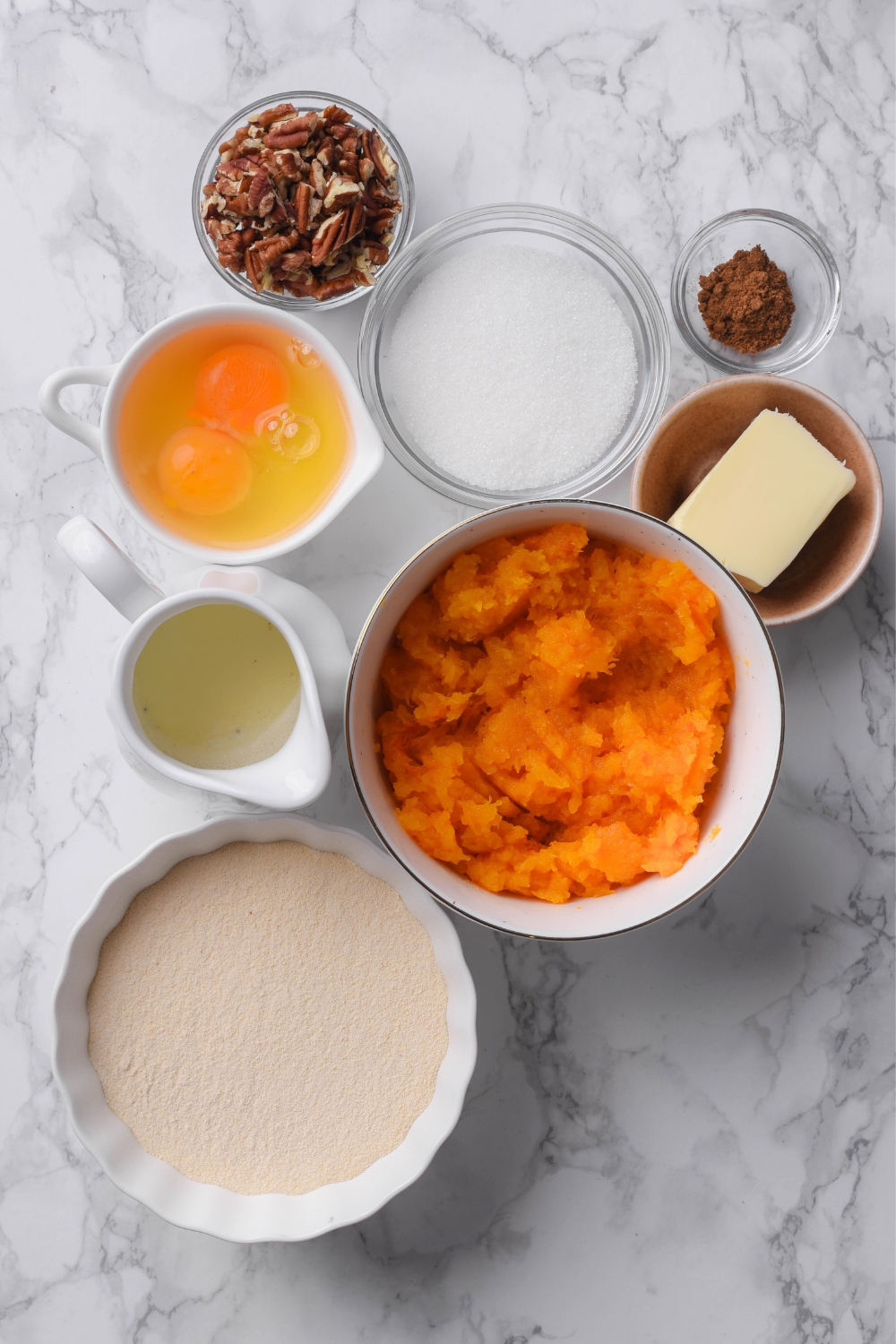 A countertop with multiple separate bowls with pumpkin puree, cake mix, butter, pumpkin pie spice, pecans, eggs, sugar, and oil.