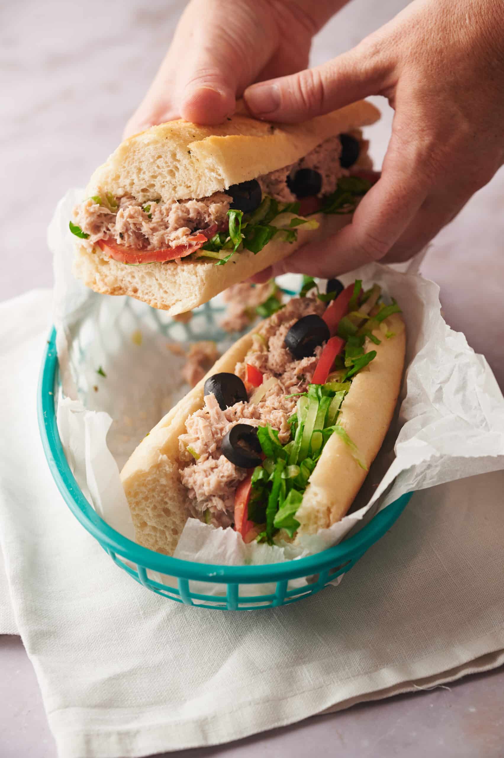 Two hands holding one half of a tuna salad sandwich. The other half is in a blue basket lined with paper.