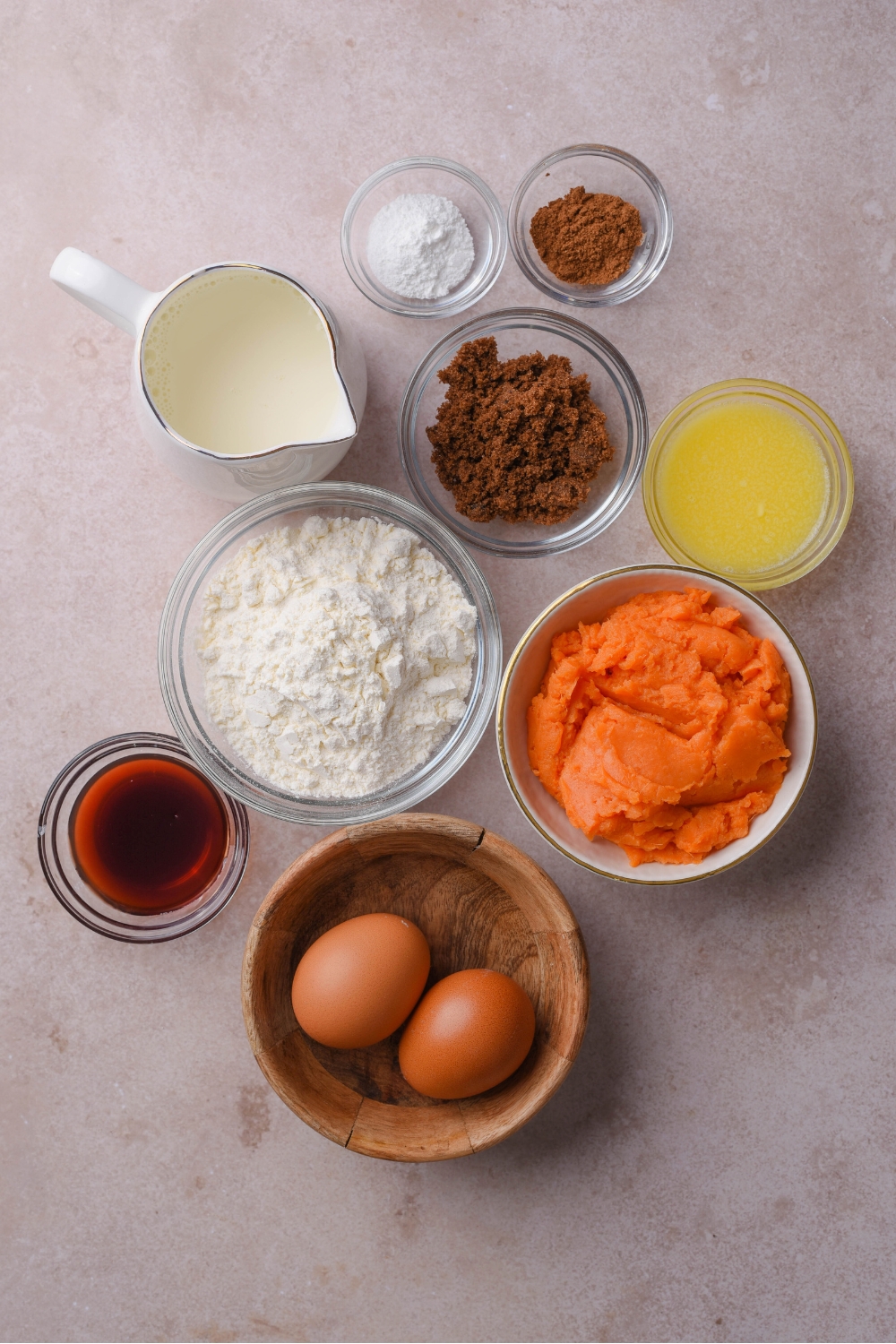 A countertop with multiple bowls with flour, mashed sweet potatoes, eggs, melted butter, maple syrup, gingerbread spices, milk, and baking powder.