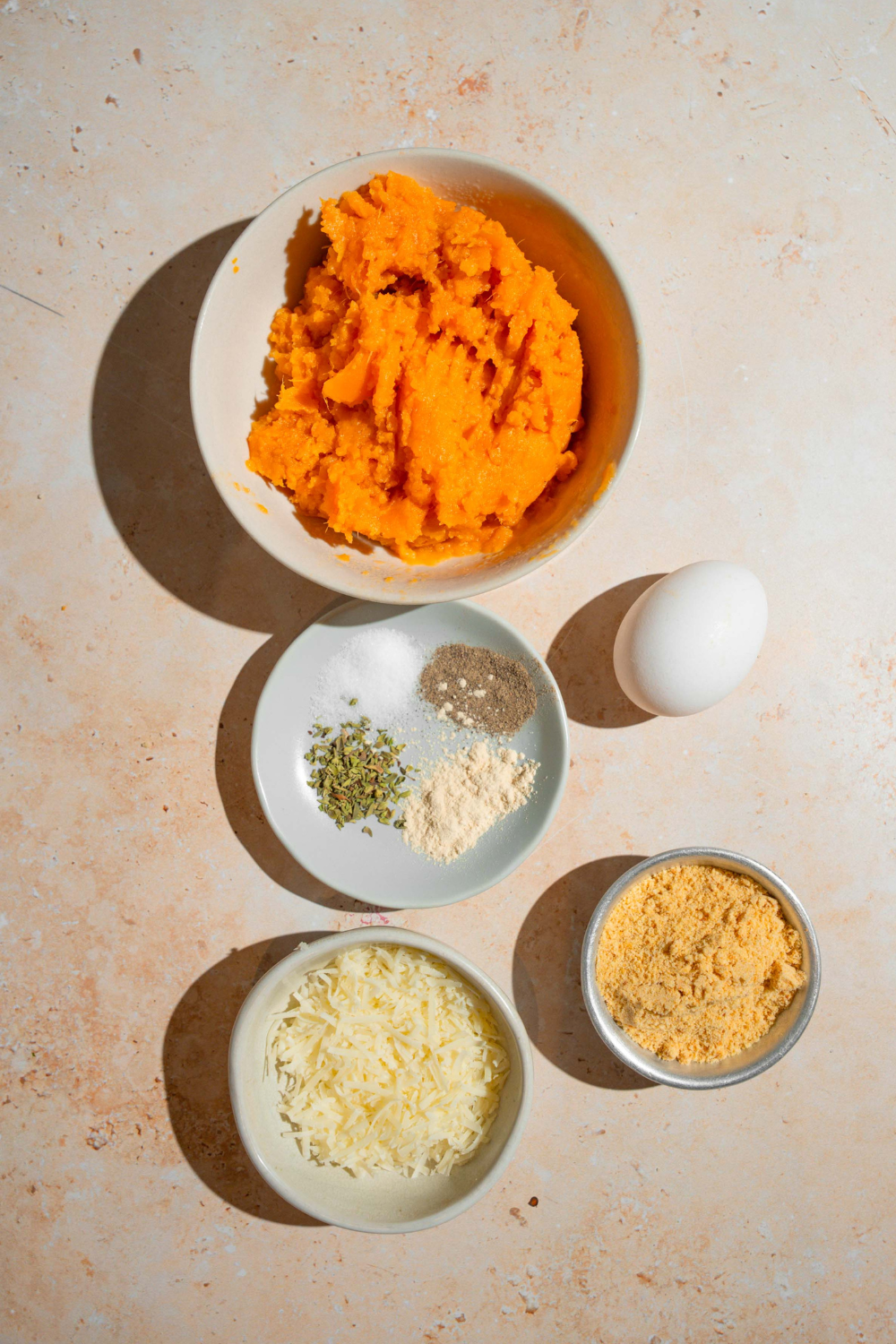 A bowl of mash, sweet potato, an egg, a small plate of seasonings, a small bowl of breadcrumbs and a small bowl of parmesan cheese all on a white counter.
