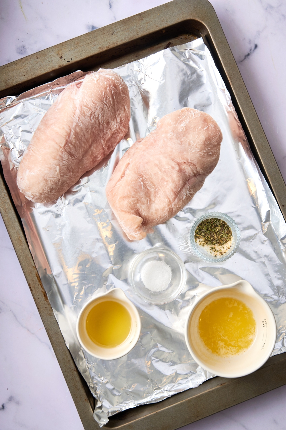Two frozen chicken breasts and small cups full of seasonings, butter, and oil are on a foil lined baking sheet.