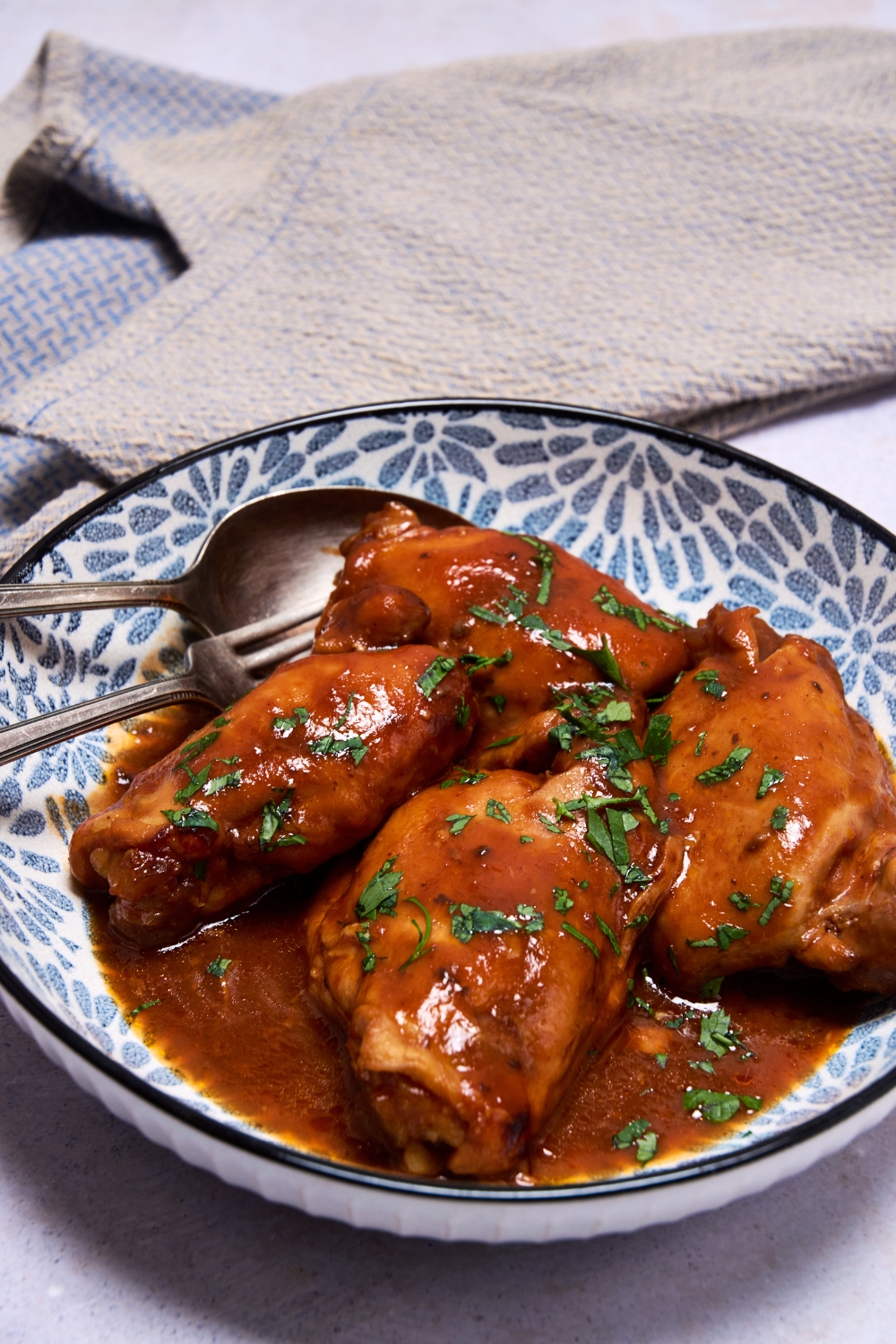 Cooked chicken thighs are in a blue and white dish. They're covered in barbecue sauce. A fork and spoon are in the dish.