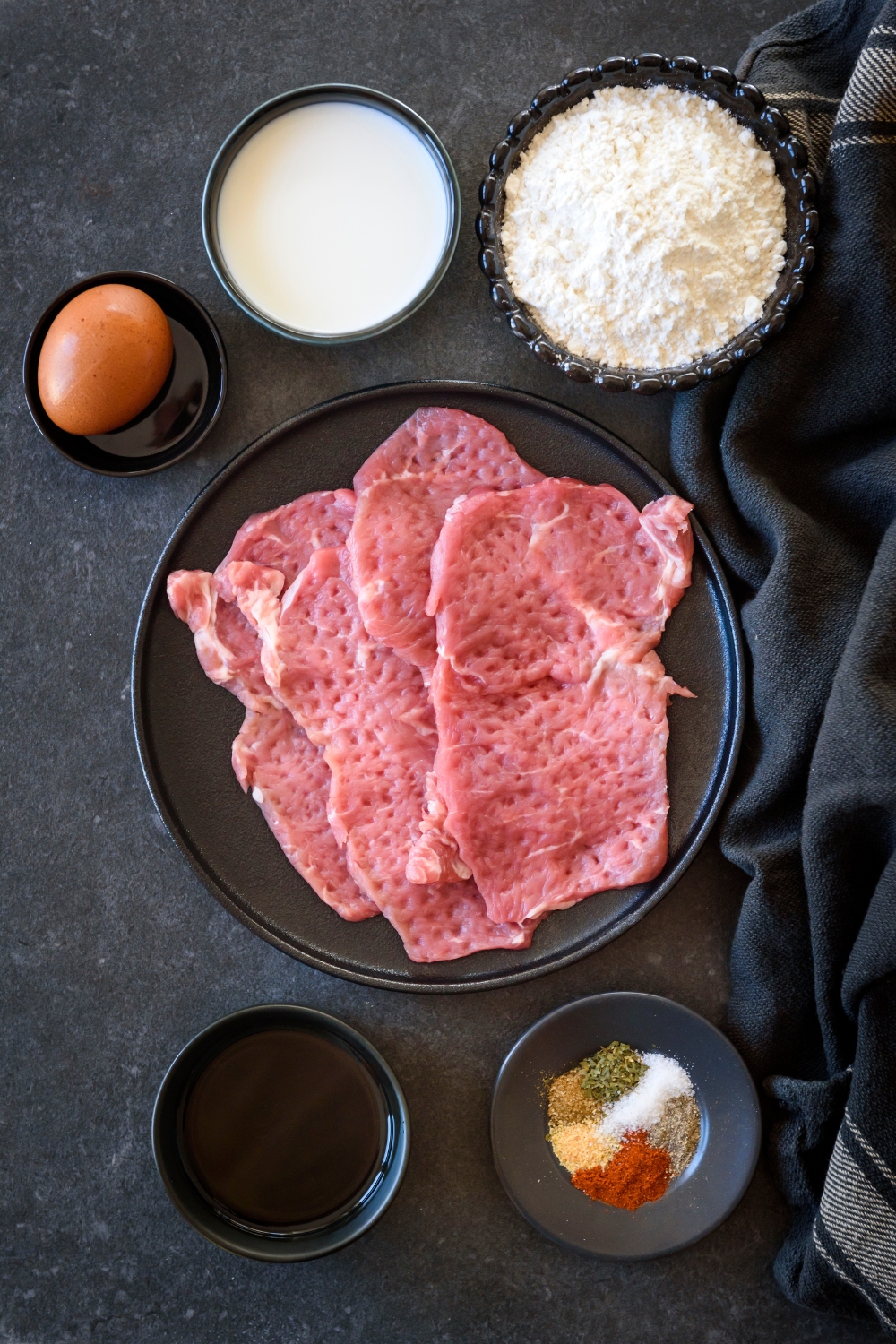 Tenderized cube steaks sit on a black plate. Flour, milk, an egg, seasonings, and vegetable oil are in separate bowls surrounding the plate.