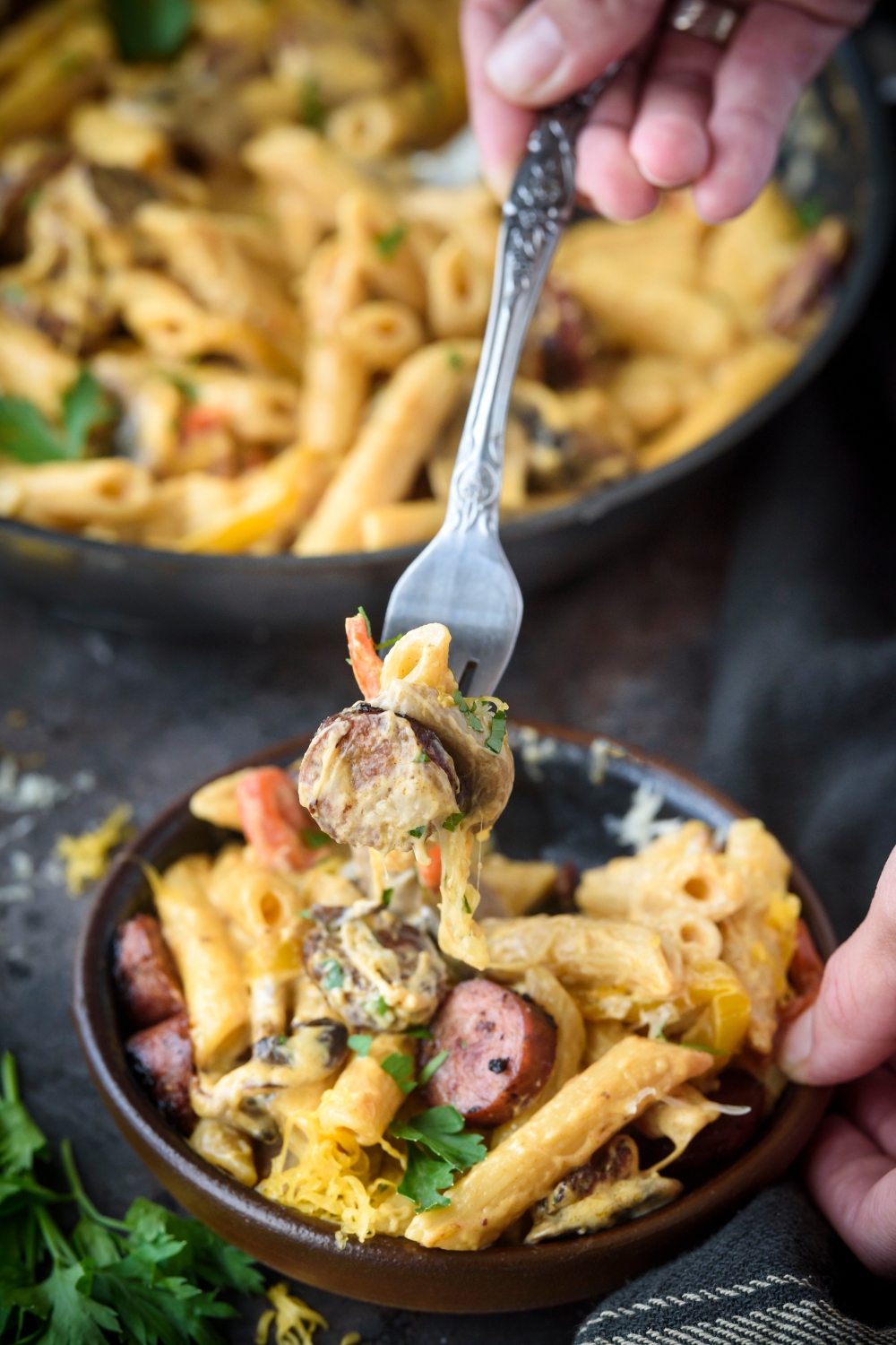 A fork holding a bite of creamy pasta with sausages. The fork is held above a bowl filled with creamy sausage pasta.