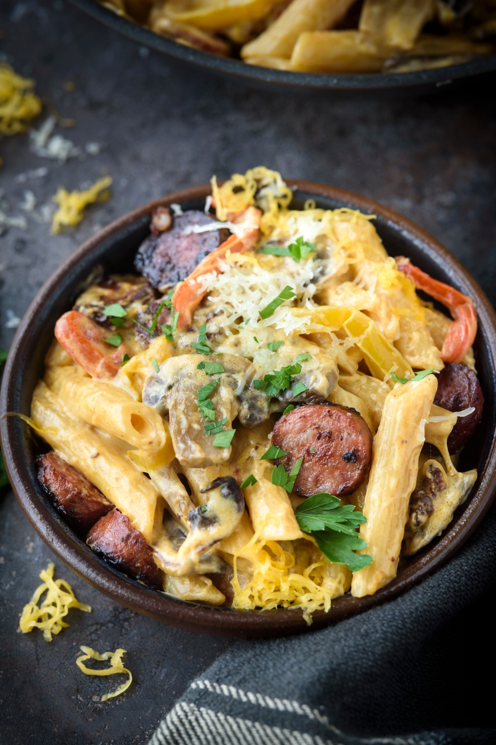 A bowl of pasta in a seasoned cream sauce with sliced sausages, red peppers, yellow peppers, shredded cheese, and fresh herbs on top.