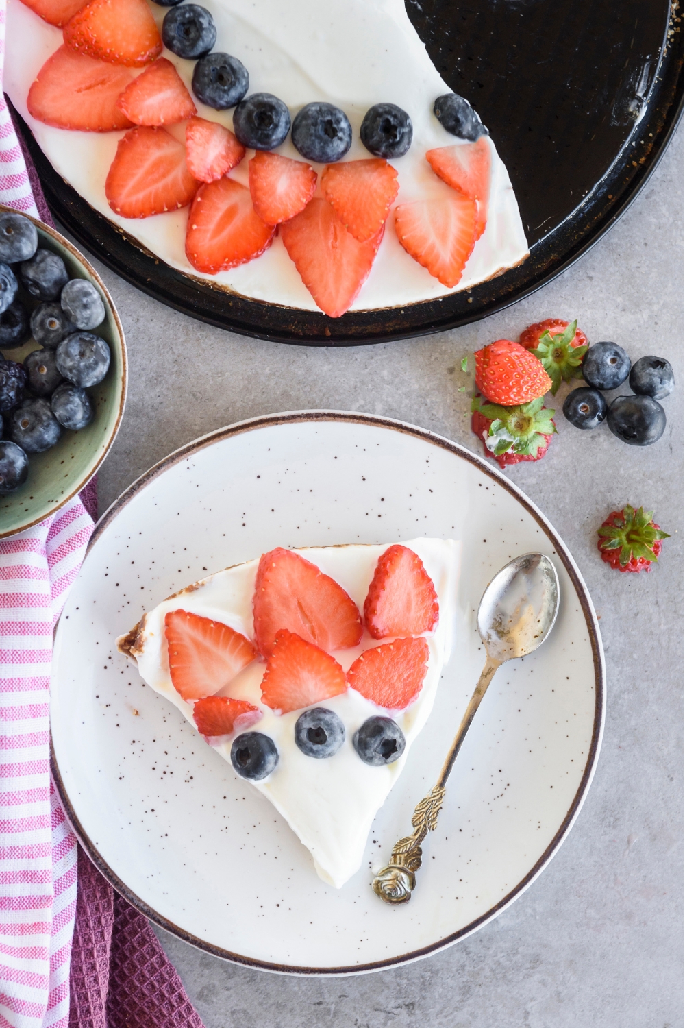 A slice of Cool Whip pie decorated with blueberries and strawberry slices. There is a spoon on the plate and the rest of the pie is in a pie pan next to the plate.