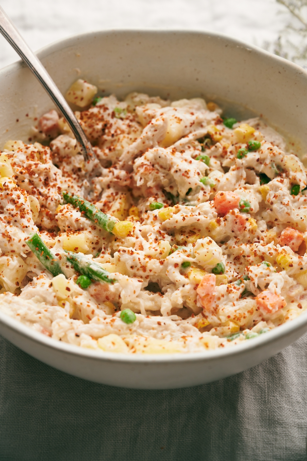 A white mixing bowl full of Mexican chicken salad sits on a gray dish cloth. A serving spoon rests in the bowl.