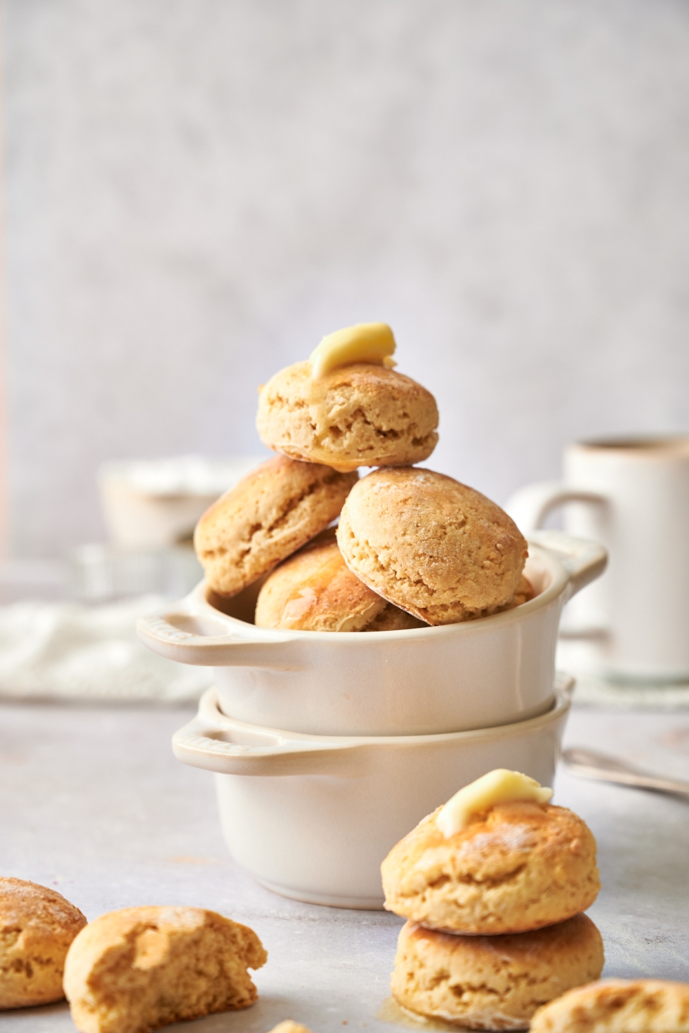 Two ramekins stand stacked on top of each other. The top ramekin is full of golden brown biscuits. The topmost biscuit has a pat of butter on top of it.