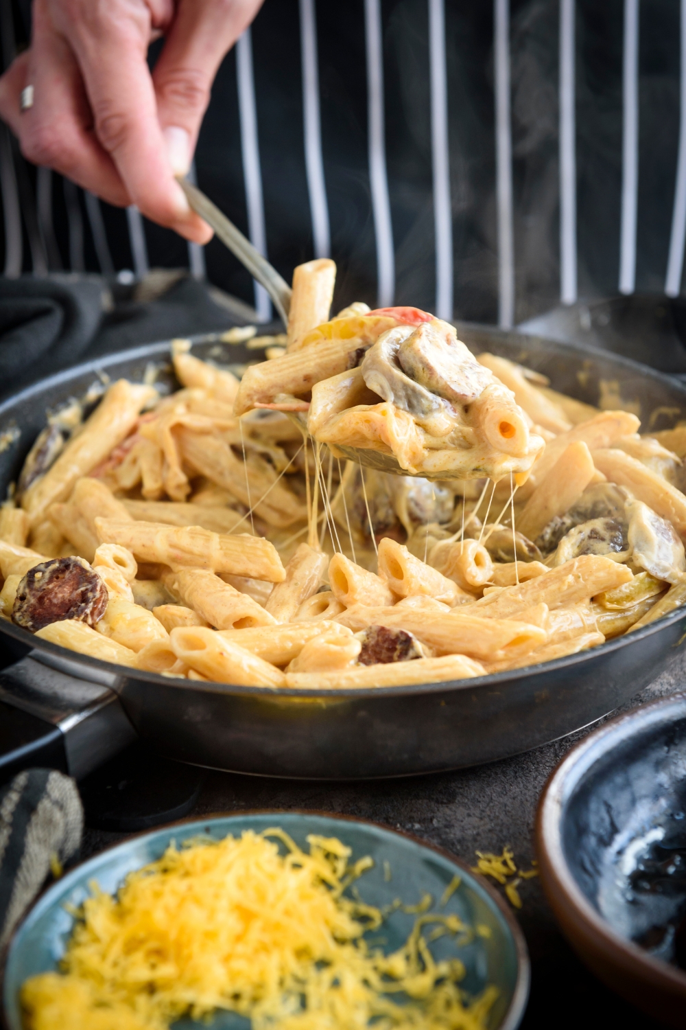 A hand scooping a spoonful of cheesy pasta mixed with peppers and sliced sausages.