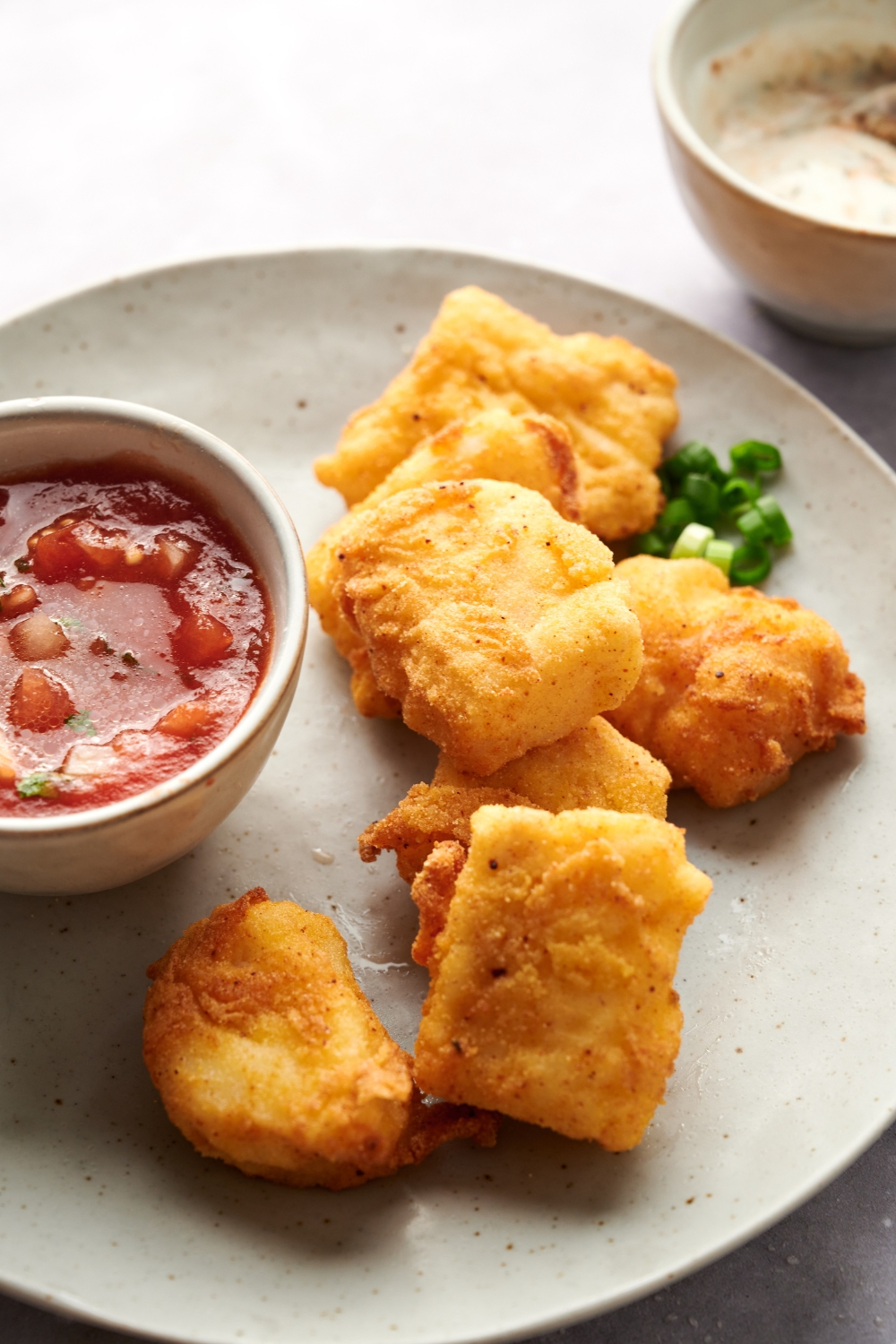 Golden brown catfish nuggets on a serving plate with a bowl of dipping sauce and chopped chives.
