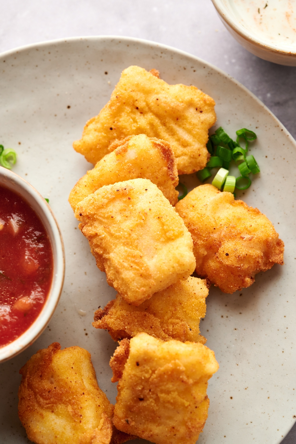 Golden brown catfish nuggets on a serving plate with a small bowl of red dipping sauce.